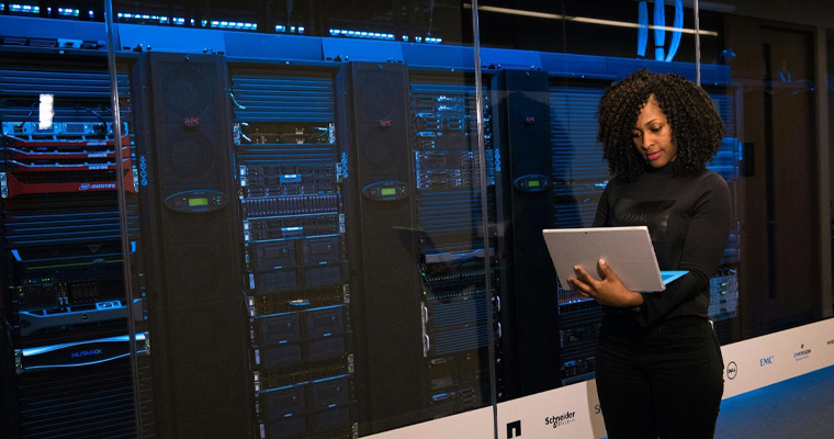 A software engineer with a laptop standing beside server racks.