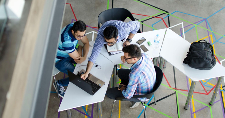 A man points at a laptop while two other men look on at a meeting. 