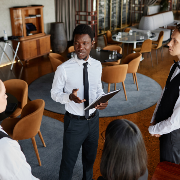 A man in formal attire, wearing a white shirt and black tie, is holding a tablet and speaking to a group