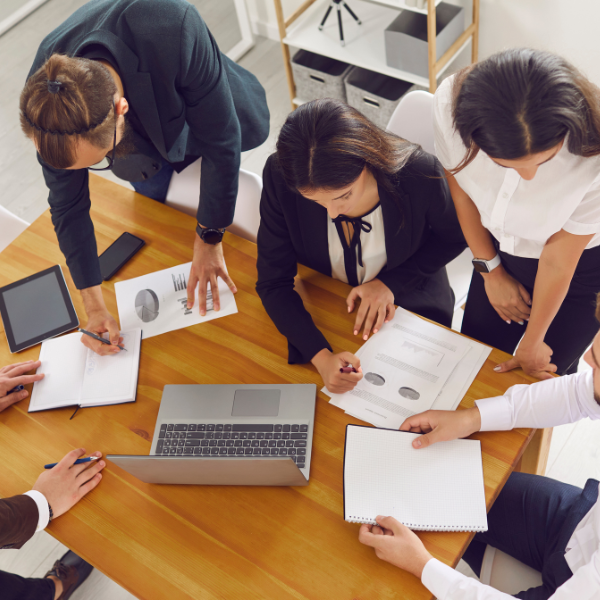 A group of people reviews documents around a table with a laptop and tablet.