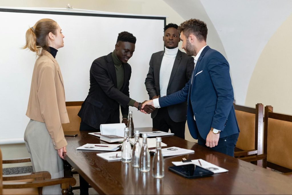 Two men shake hands at a conference table while two other people look on.