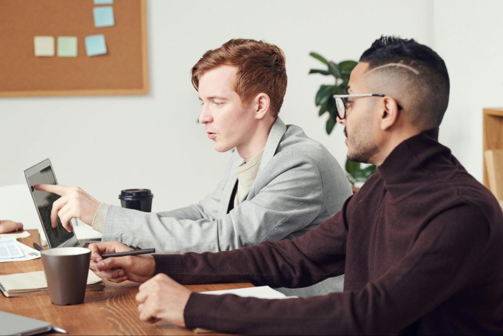 A man points at a laptop on a desk while another man looks on.